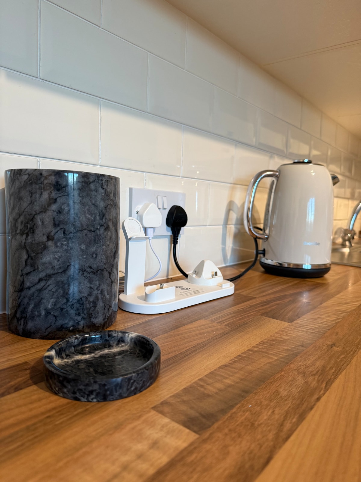 A kitchen countertop is showcased with a polished wooden surface. A sleek electric kettle is positioned alongside a marble canister and a small dish. A charging station is visible, featuring multiple outlets and space for devices.