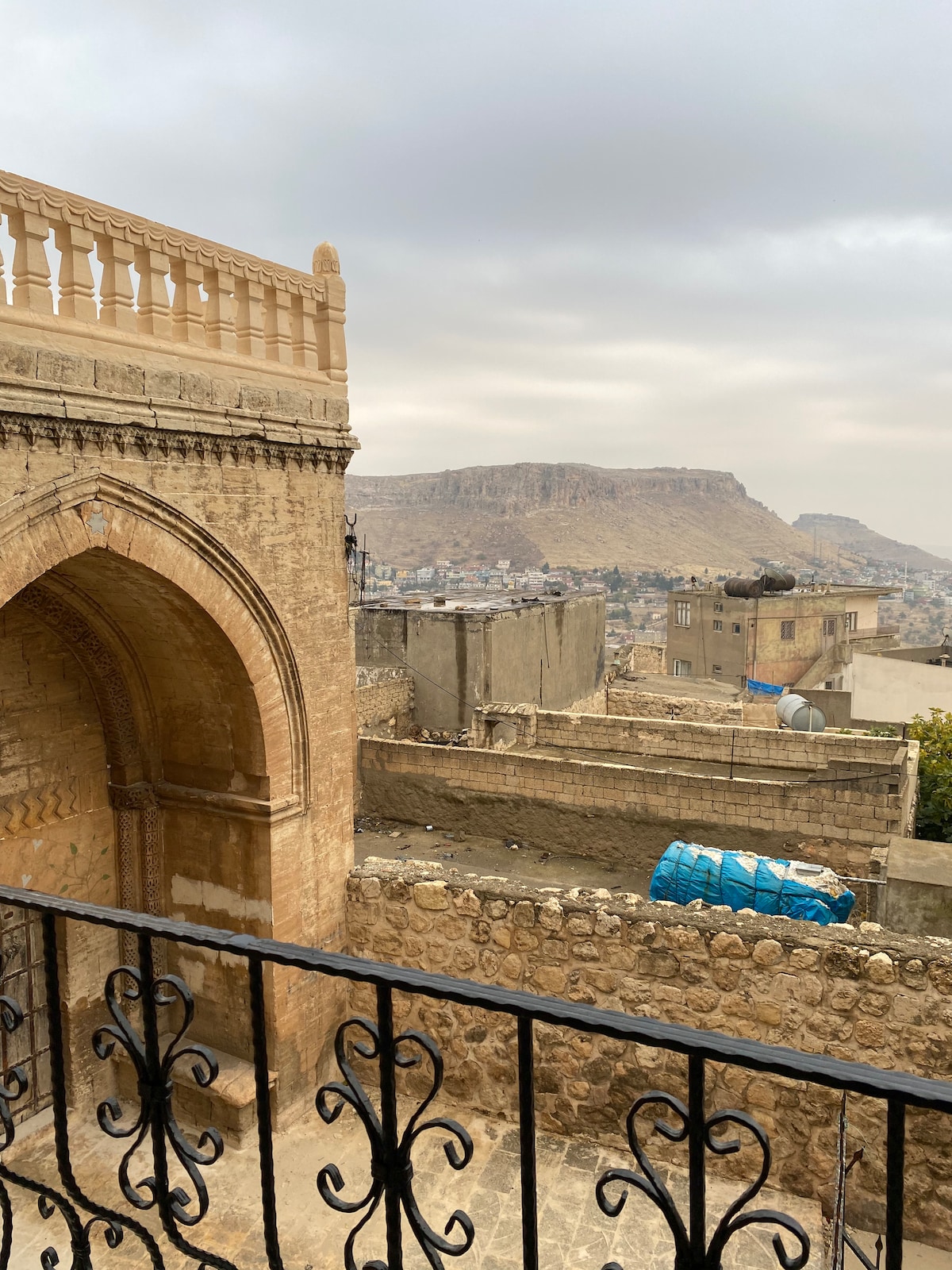 An arched stone structure is visible in the foreground, leading to a balcony with decorative iron railings. The backdrop features a mountainous landscape under a cloudy sky, with various buildings and a blue water tank in the distance.