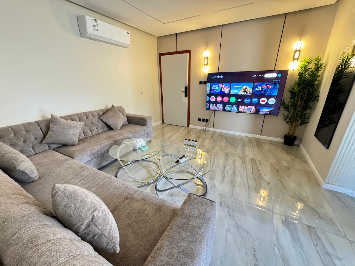 A modern living area is presented with a plush sectional sofa, accented by decorative pillows. A sleek glass coffee table sits in front, with a smart TV mounted on the wall showcasing vibrant content. Potted plants add a touch of greenery, and natural light enhances the space.