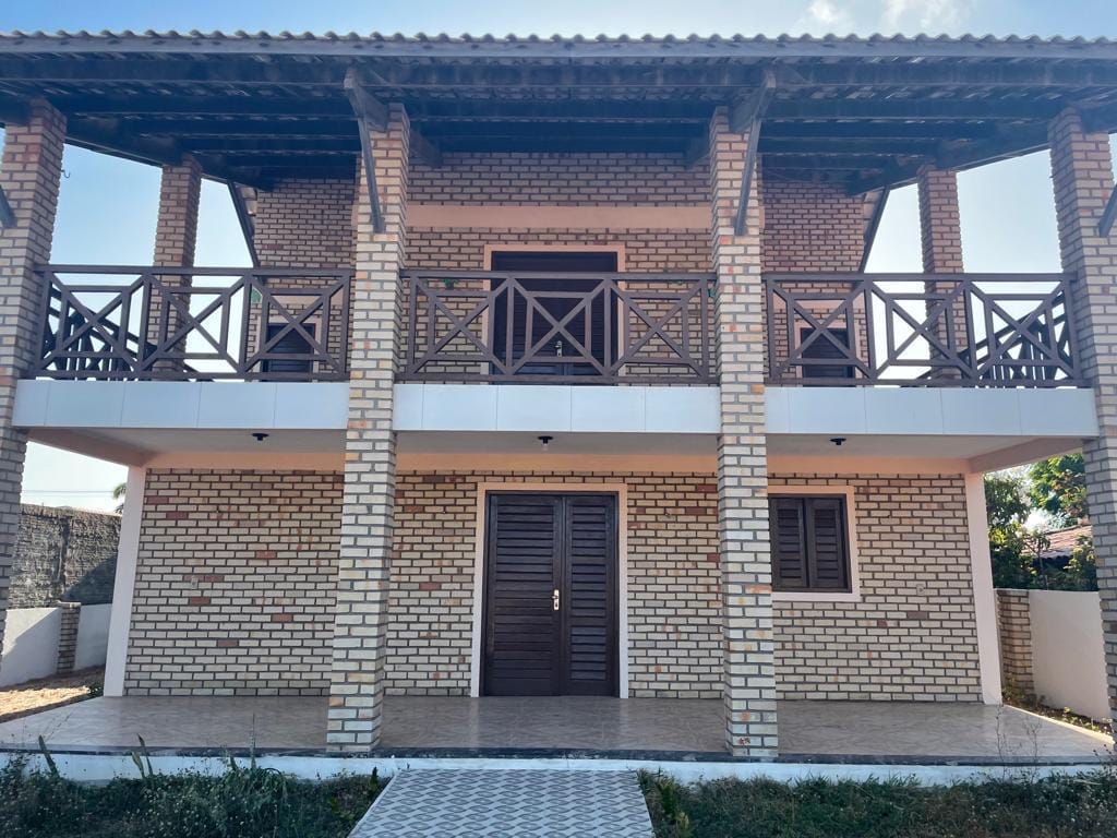 A two-story duplex house is displayed, featuring a modern brick facade and ample balcony railings. The entrance is framed by double doors, with windows on either side. A tiled pathway leads to the front, and a well-maintained garden area is seen surrounding the property.