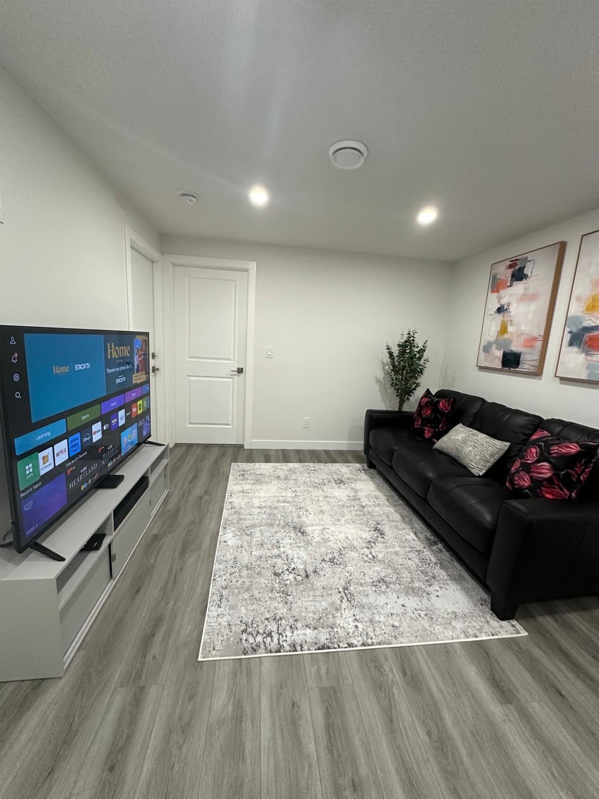 A modern living area is shown with a sleek black sofa adorned with decorative pillows. A large television is mounted on a white media console, accompanied by a soft area rug that adds warmth to the wooden floor. Bright wall art provides a pop of color.
