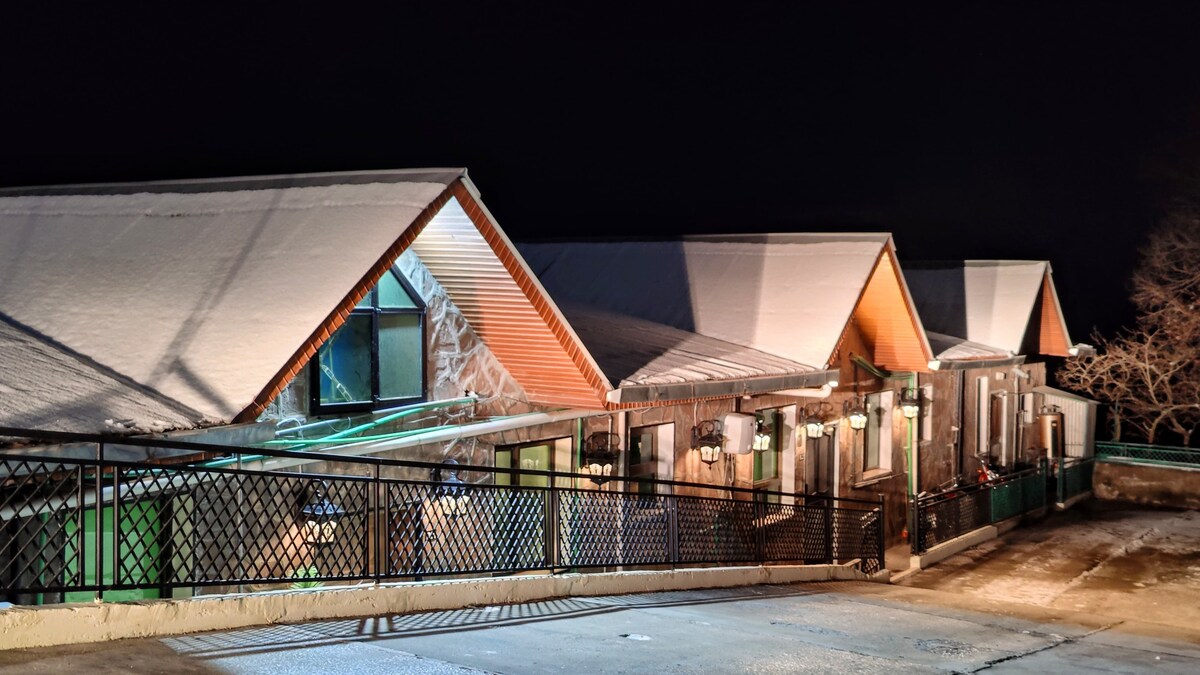 The exterior of the duplex cottage is displayed at night, featuring a snow-covered roof and sleek architectural lines. Soft lighting highlights the entrance, while the surrounding area is enclosed by a decorative black railing, enhancing the property's elegance and secure ambiance.