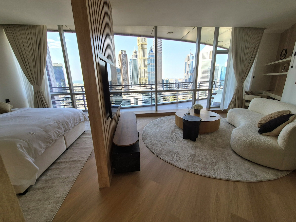 An open living space is displayed, featuring a spacious bed adjacent to a minimalist seating area. Large windows afford views of the skyline. A round coffee table and a soft, round sofa complement the neutral tones of the room, while light pours in through sheer curtains.