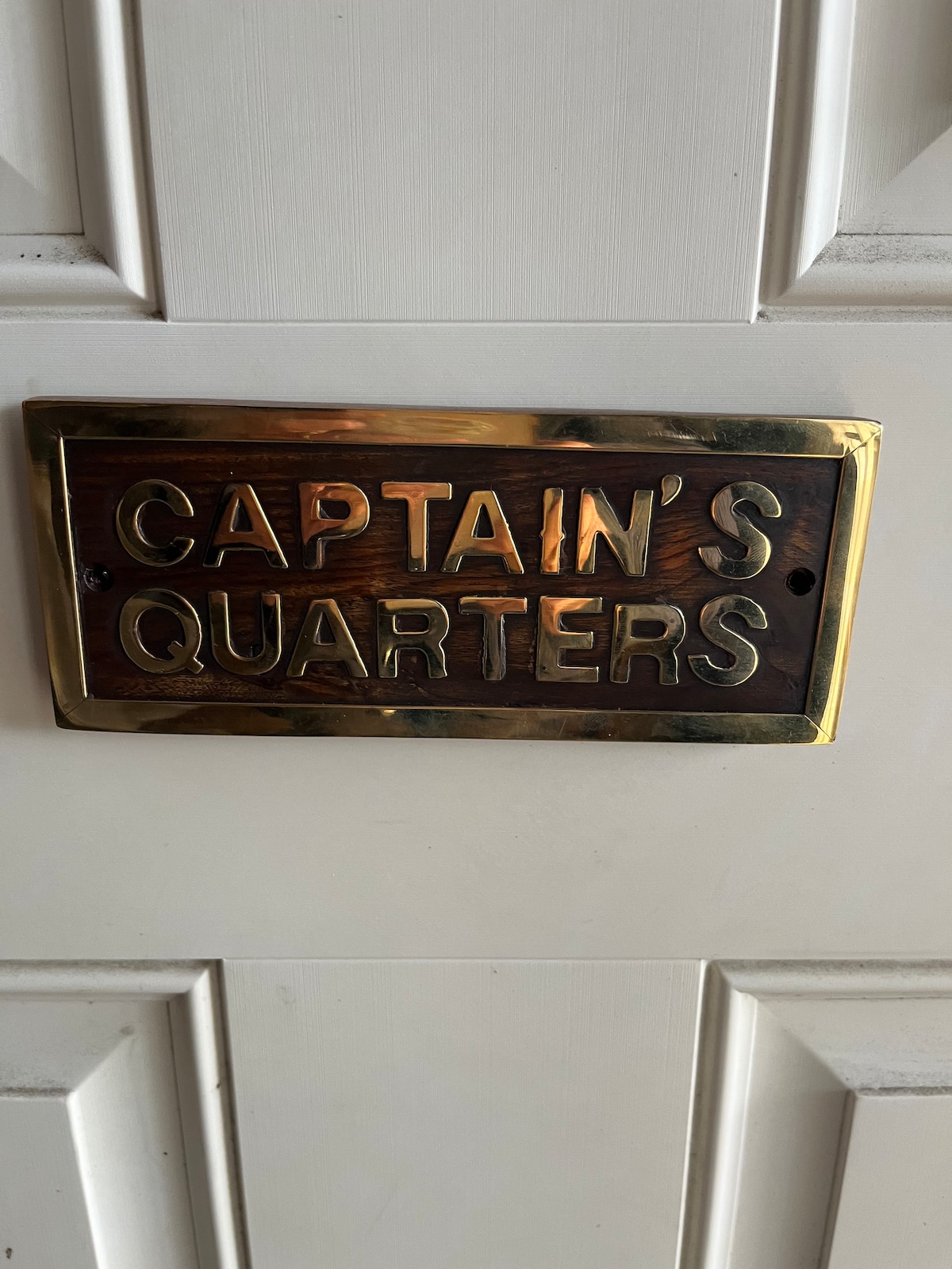 A brass plaque labeled 'CAPTAIN'S QUARTERS' is mounted on a wooden door. The plaque's lettering stands out against a rich background, suggesting a nautical theme. Light reflections can be seen on the polished surface, enhancing its visibility.