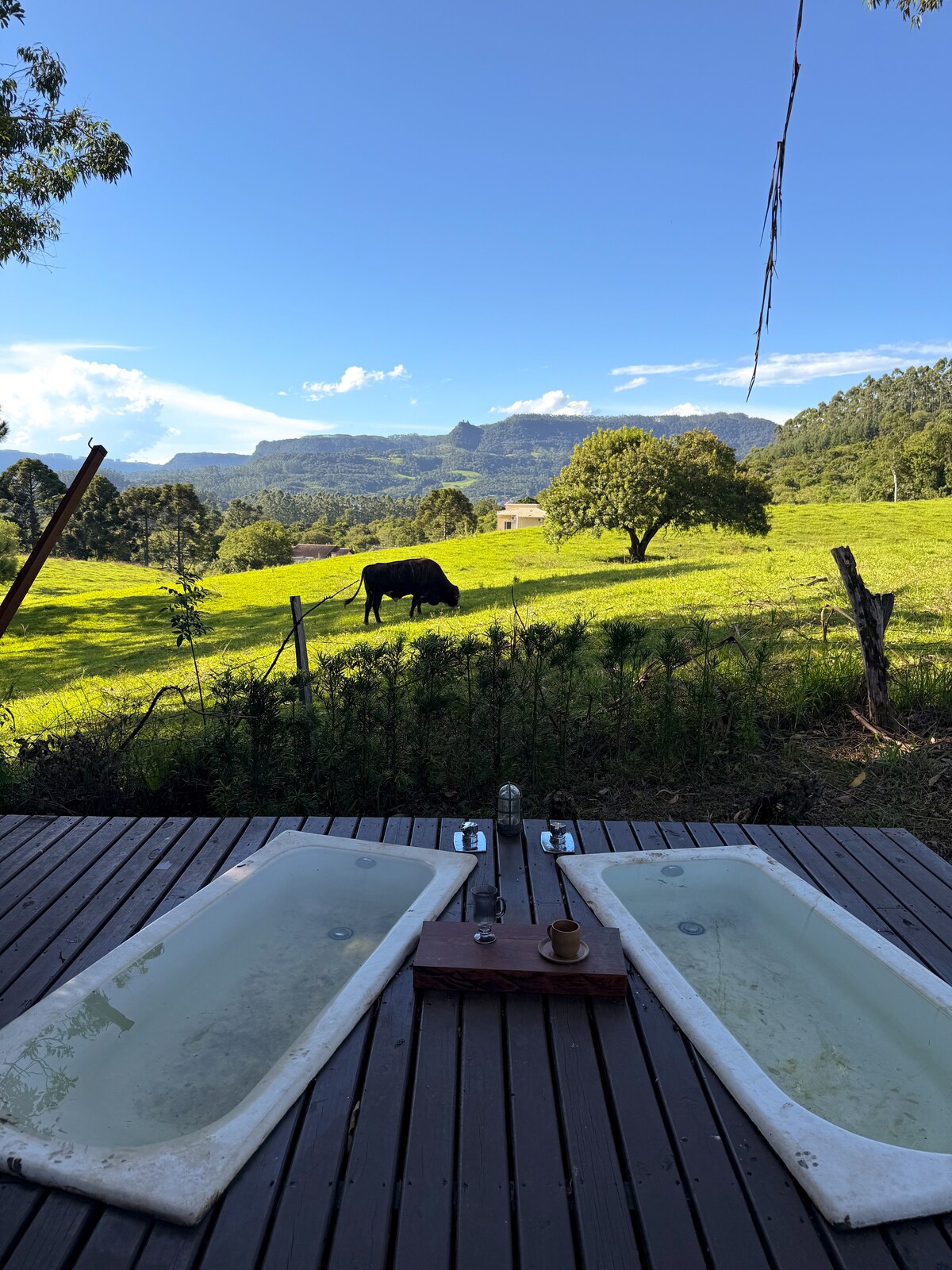 Two outdoor bathtubs are positioned on a wooden deck, offering a view of a green landscape. A cow grazes in the distance, with rolling hills visible under a clear blue sky and scattered clouds.