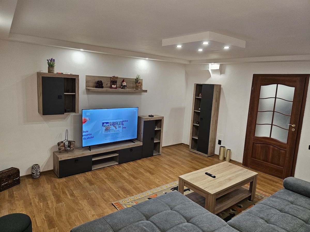 A spacious living room is presented with a large flat-screen television mounted on a neutral-colored entertainment unit. A coffee table sits in front of a comfortable gray sofa, with wooden flooring and soft lighting enhancing the room's inviting appearance. A decorative rug adds warmth to the space.