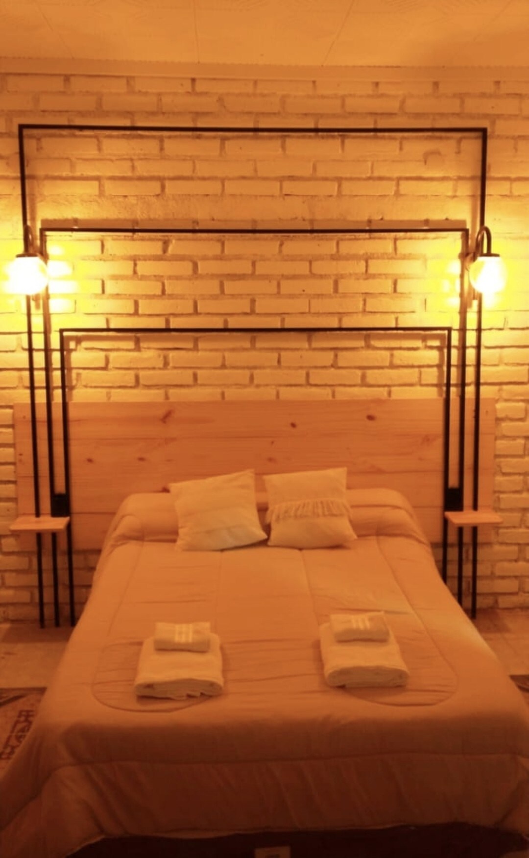 A cozy sleeping area features a wooden headboard and soft bedding. Two bedside tables with warm lighting fixtures flank the bed, providing a soft glow against the textured brick wall. Neatly arranged towels lie on the bed, enhancing the welcoming appearance of the space.