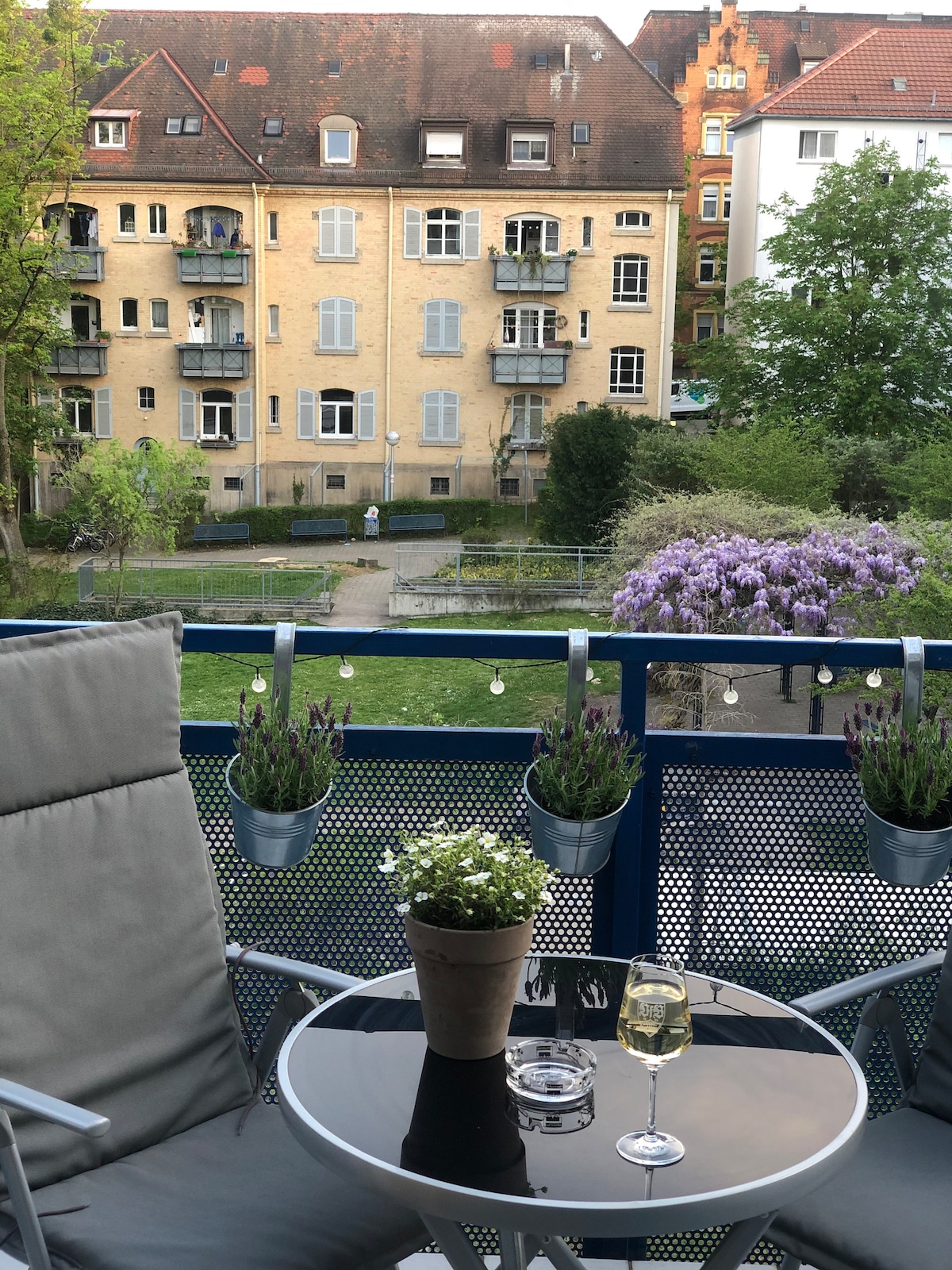 A balcony view reveals a peaceful courtyard adorned with greenery and flowering plants. A round table holds a glass and a potted plant, while hanging planters add to the serene ambiance. The backdrop features a charming yellow building with multiple balconies.