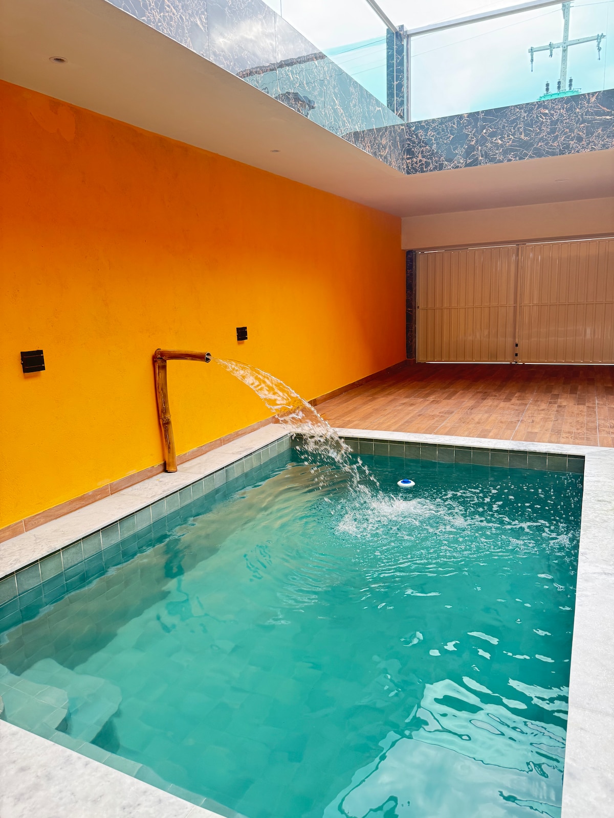 A compact pool area features turquoise water with a gentle waterfall creating a soothing sound. The vibrant orange wall adds a lively touch, while a covered section offers protection from the elements. The surrounding floor is composed of wooden tiles, providing a natural warmth.