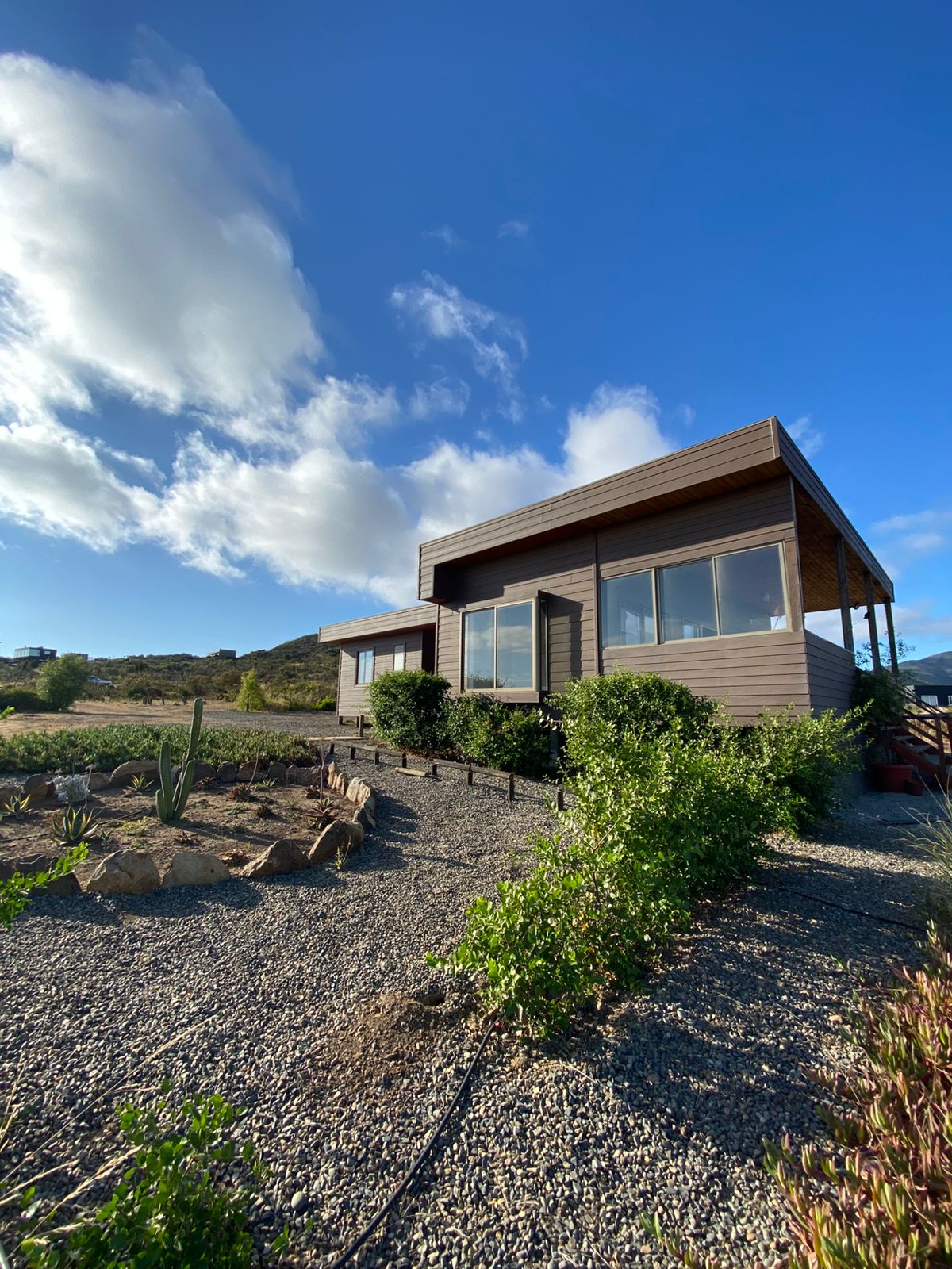 A modern house is pictured with a unique architectural design, surrounded by native vegetation and gravel pathways. Large windows allow for ample natural light, framing the scenic views of the landscape and sky. The structure sits elevated, providing an inviting presence.