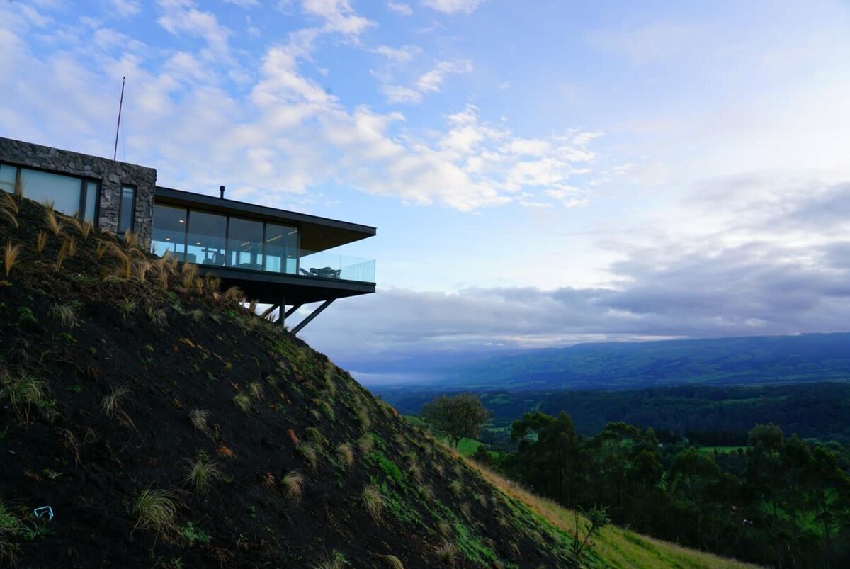 A modern structure is positioned on a hillside, featuring large glass windows that provide expansive views of the surrounding landscape. The building's design incorporates natural stone elements, and a deck extends outward, allowing for outdoor enjoyment of the panoramic scenery.