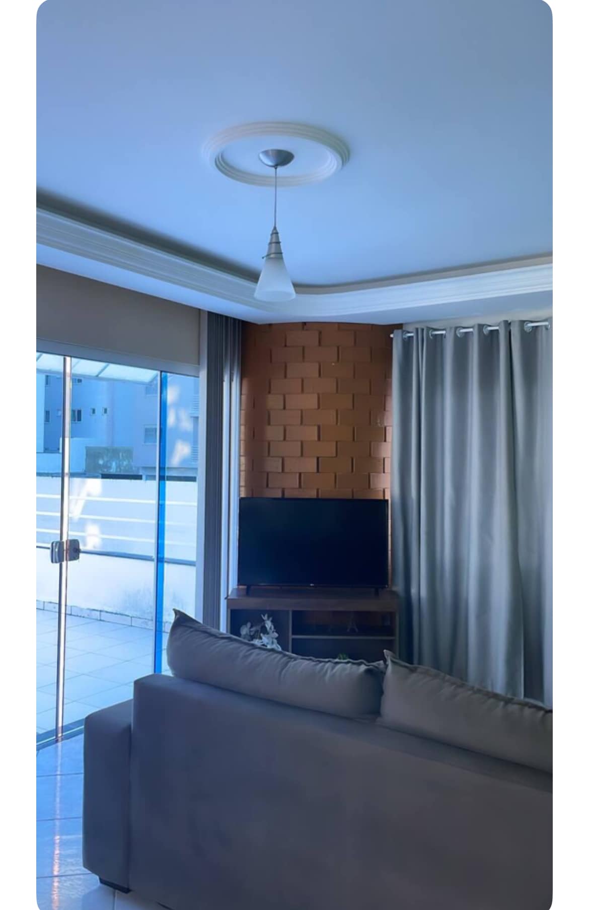 A well-lit living area features a modern television placed on a wooden unit against a textured brick wall. Large sliding glass doors invite natural light, while soft gray curtains frame the windows, enhancing the spacious feel of the room.