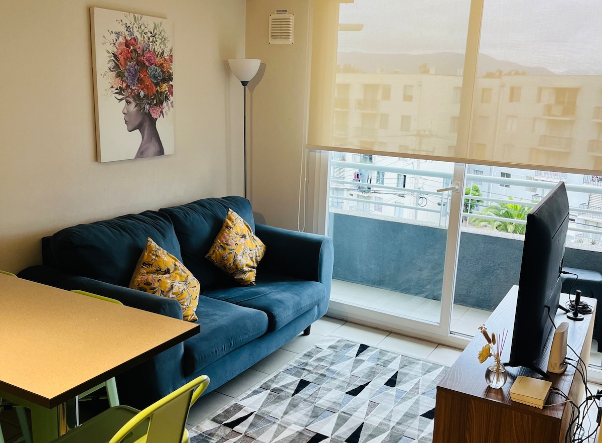 A cozy living area is highlighted by a blue sofa adorned with decorative pillows. A flat-screen TV is positioned on a wooden unit, while a dining table with colorful chairs is featured. A large window allows natural light to fill the room, enhancing the open feel.