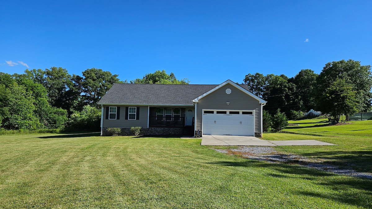 The home is set on a sprawling green lawn, surrounded by trees. The exterior features a combination of stone and siding, with a covered porch inviting guests. The driveway leads to a two-car garage, complementing the spacious and serene landscape.