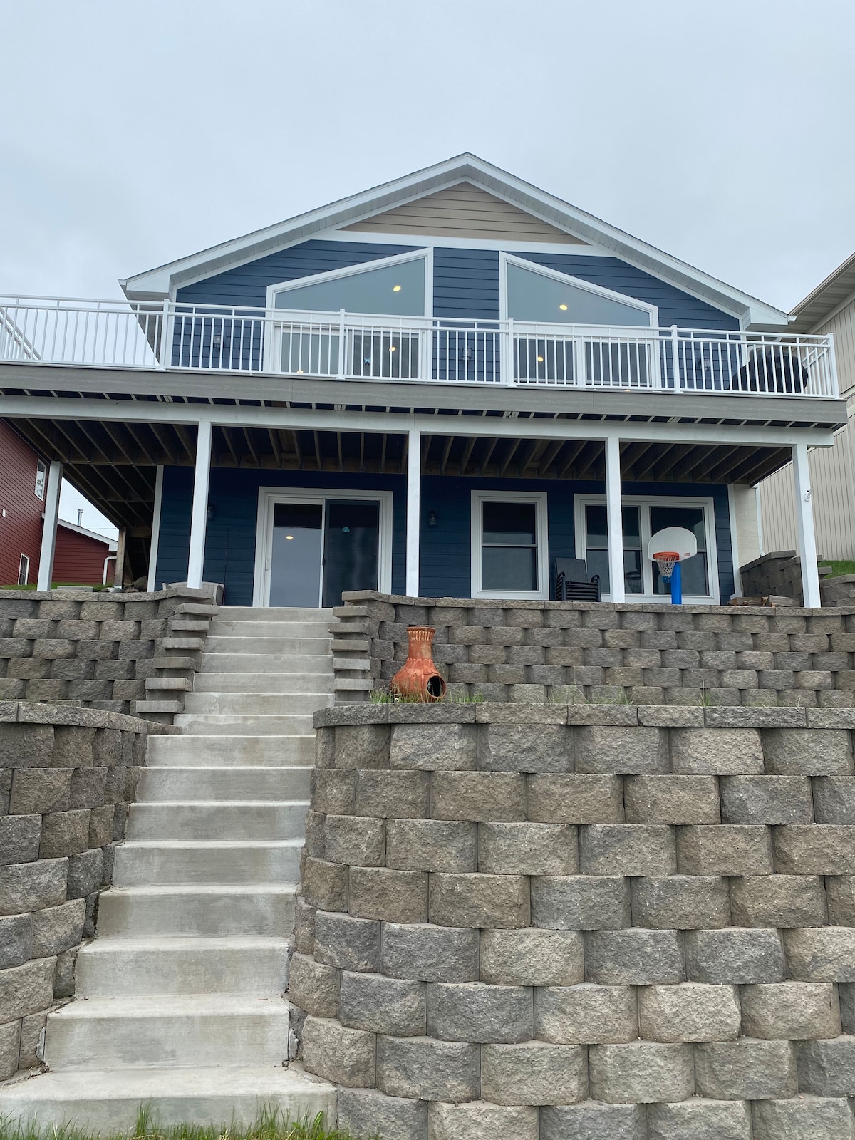 The exterior of the blue bungalow showcases a tiered retaining wall leading to a concrete staircase. A spacious deck is visible on the upper level, while large glass doors provide access to the main entrance. A basketball hoop is positioned nearby.