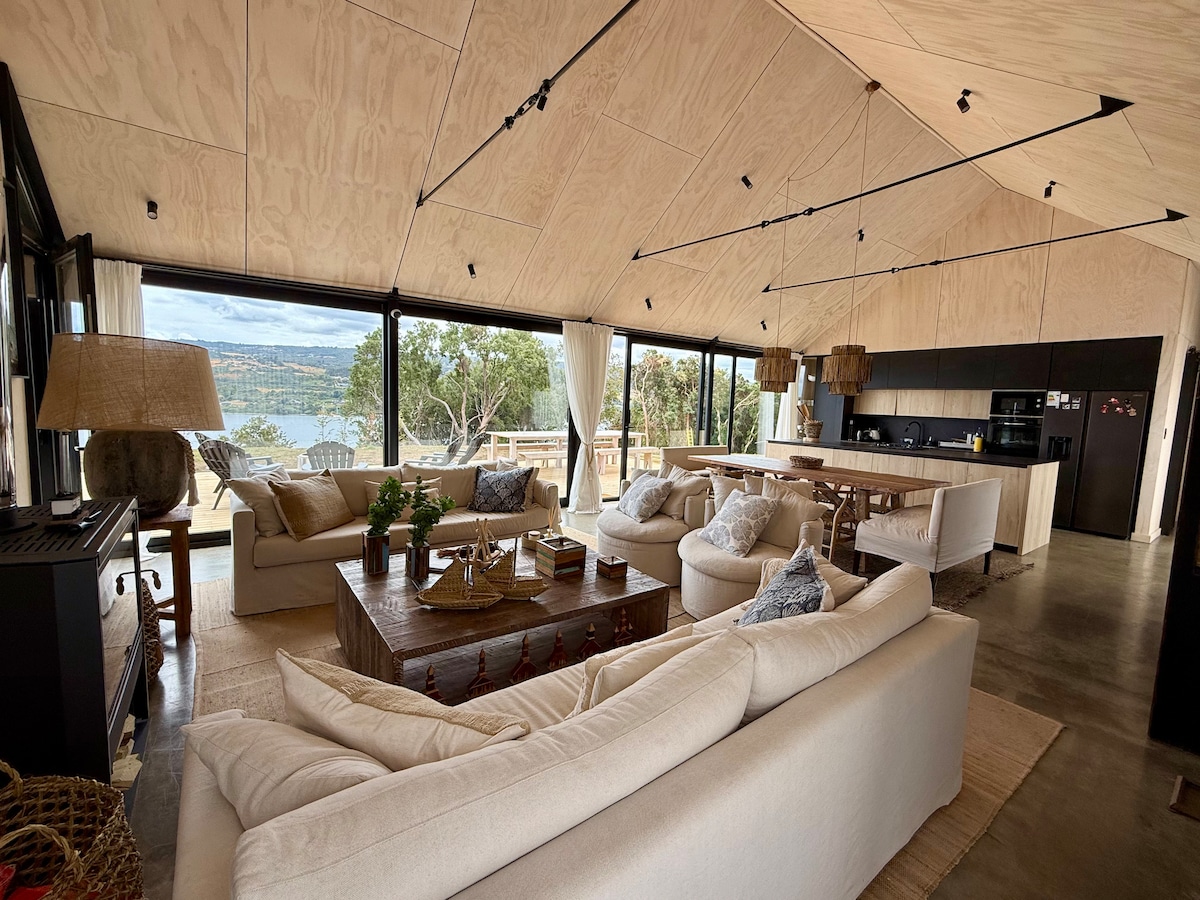 An open living area features large floor-to-ceiling windows that provide views of the water and surrounding landscape. A spacious arrangement of light-colored sofas encircles a wooden coffee table. In the background, a modern kitchen with an island is visible, enhancing the communal atmosphere.