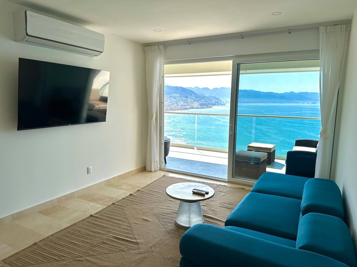 A modern living area features a deep blue couch facing a wall-mounted television. Large glass doors open to a balcony with ocean views, allowing natural light to fill the space. A round coffee table sits on a textured area rug, creating a comfortable gathering spot.