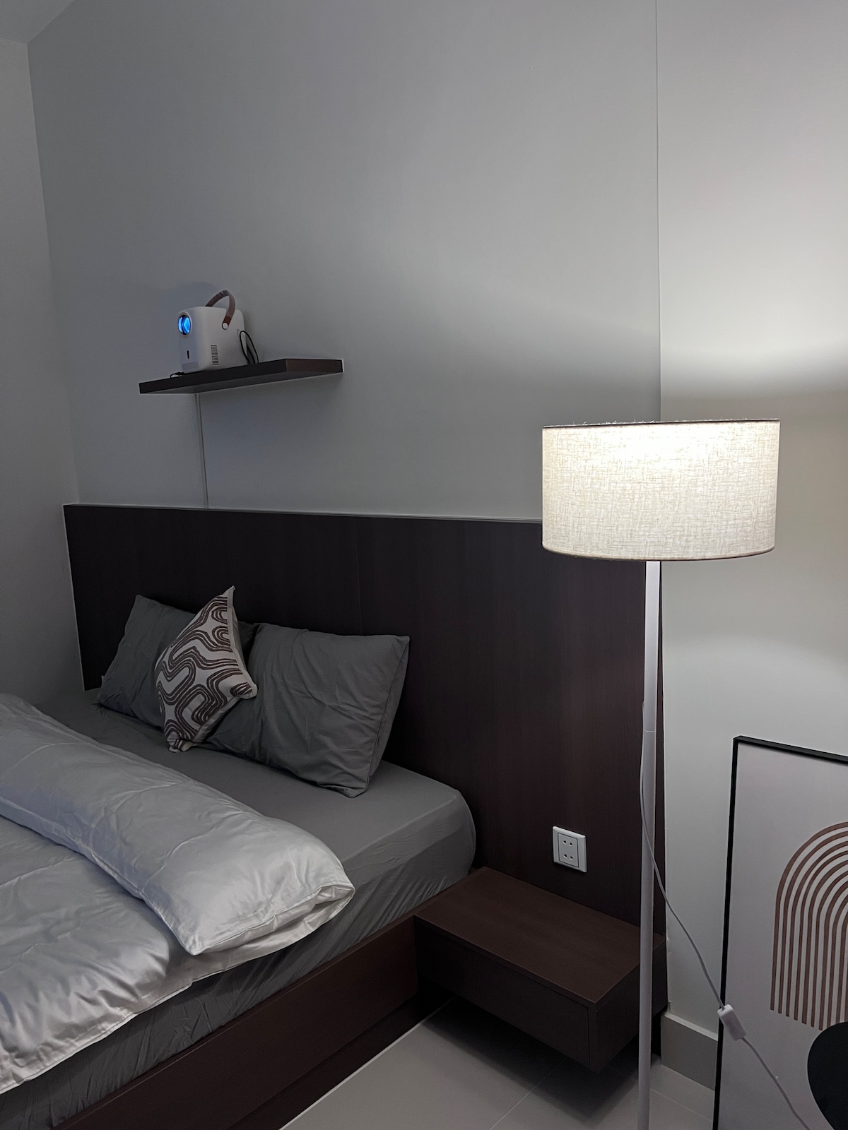 A cozy bedroom features a spacious bed adorned with neutral linens and a decorative pillow. A bedside table is positioned next to the bed, with a modern lamp providing soft lighting. A simple shelf above the bed displays minimal decor, maintaining a clean aesthetic.