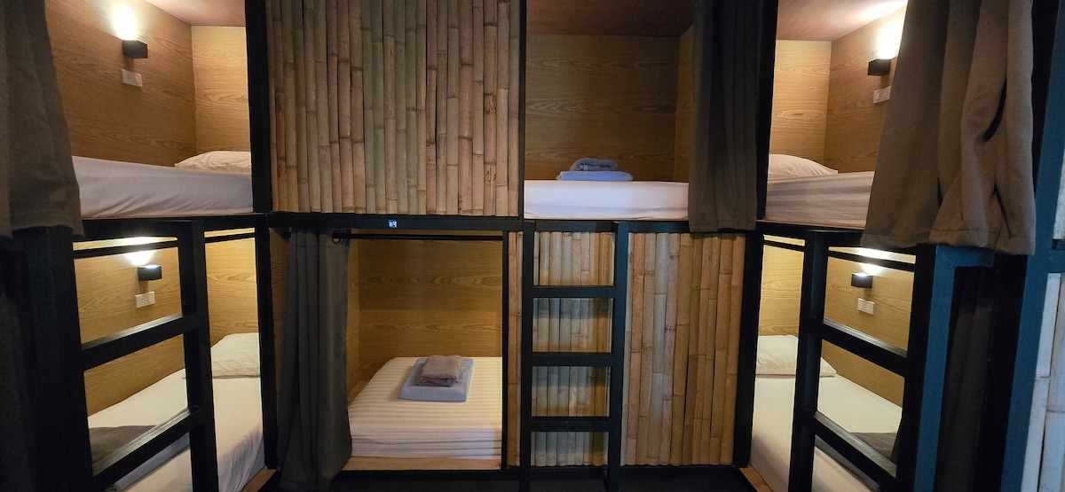 A shared dormitory space features four capsule beds, each framed by natural bamboo walls. Soft lighting is positioned above each bed, creating an intimate ambiance. Neutral-colored linens and towels are neatly arranged on the beds, while curtains provide privacy.