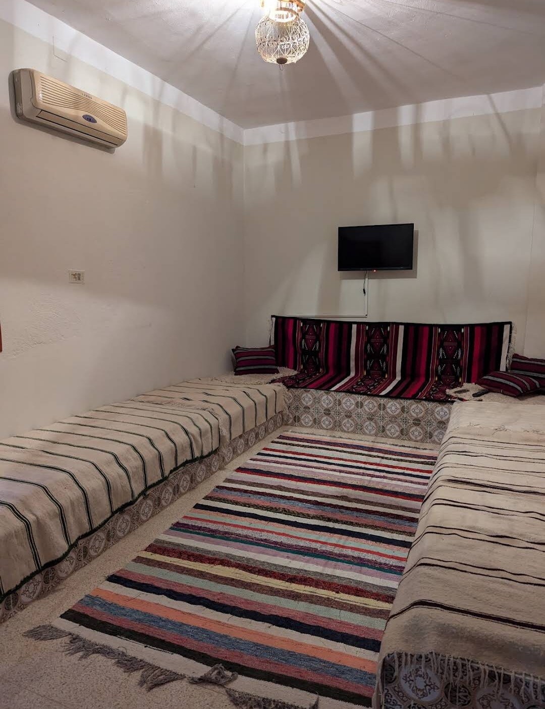 A cozy seating area features low cushions and traditional textiles in warm tones. A flat-screen television hangs on the wall across from a patterned rug that adds color to the floor. Soft light is emitted from a decorative ceiling fixture.