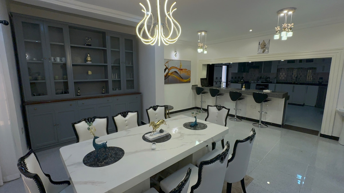 A spacious dining area is highlighted by a large, modern table surrounded by elegantly designed chairs. A stylish light fixture hangs overhead, while a sleek kitchen bar with stools is visible in the background. Decorative items are placed on the table and shelves.