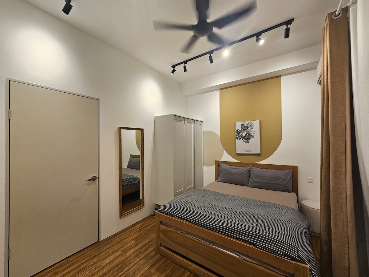 A serene bedroom features a spacious bed adorned with neutral-toned bedding. A mirror reflects the room, and a closet stands against the wall. Soft lighting highlights the simple artwork above the bed, complemented by a ceiling fan and light wooden flooring.
