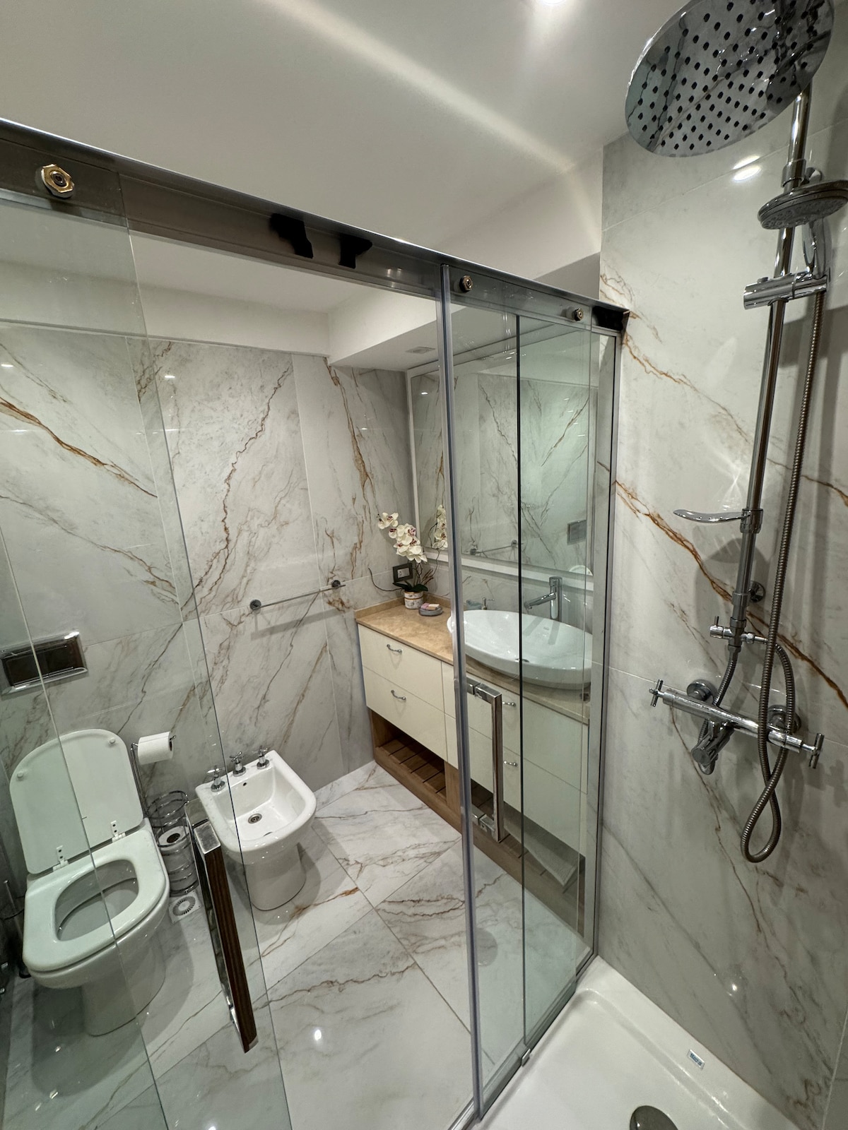 A modern bathroom is highlighted with marble walls and a sleek glass shower enclosure. A spacious vanity with an integrated sink is positioned against the wall. A bidet and a toilet are situated within the area, offering a clean and functional design.