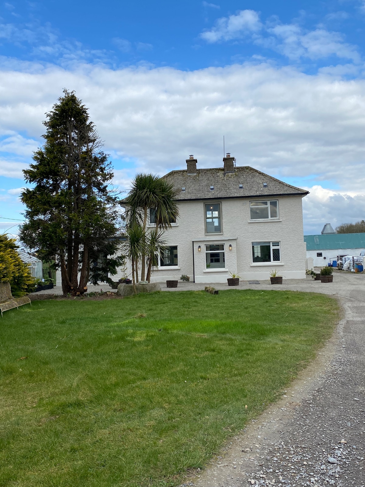 The farmhouse is a two-story white building situated on a well-kept grassy area surrounded by a variety of plants and trees. The entrance features a pathway leading to the main door, with a scenic view of the surrounding countryside in the background.