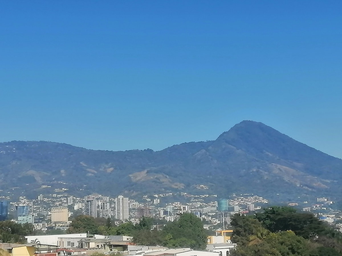 A clear view showcases a mountainous landscape against a bright blue sky, highlighting the contrast between the natural terrain and urban structures below. The city skyline features a mix of buildings, creating a vibrant backdrop to the mountainous scenery.