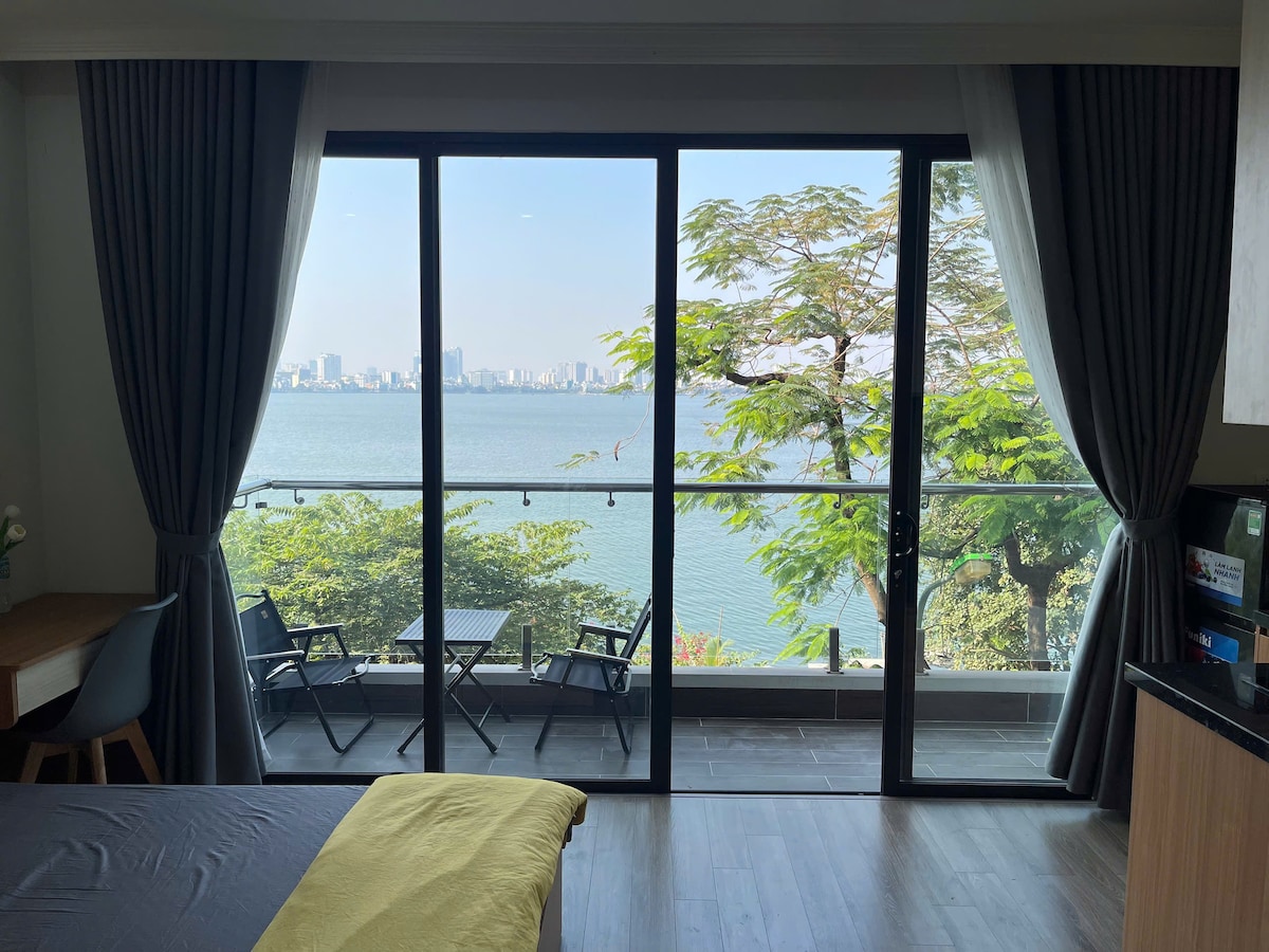 Large glass doors provide unobstructed views of the lake and city skyline, allowing natural light to fill the room. A small dining table and two chairs are positioned on the balcony, surrounded by greenery, enhancing the relaxing atmosphere.
