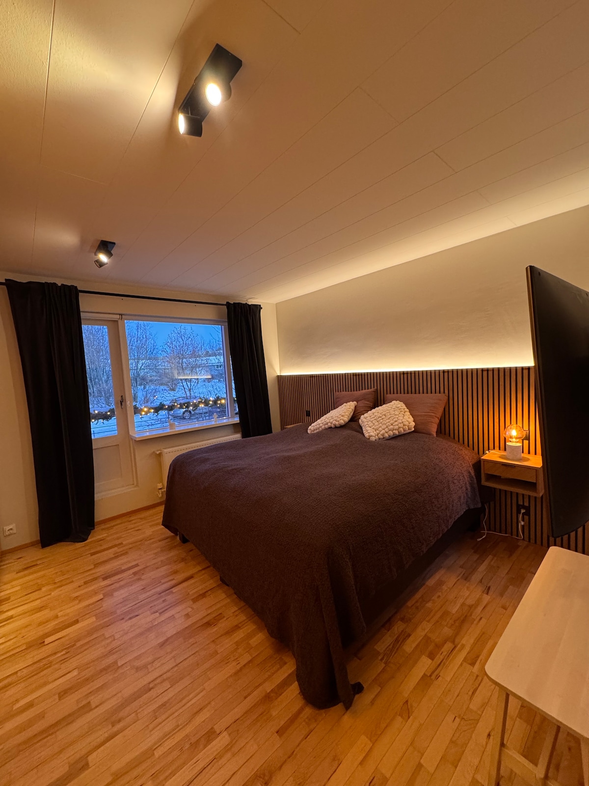 A well-lit bedroom features a spacious bed dressed in a deep brown coverlet. Soft lighting outlines the headboard, creating a gentle ambiance. Large windows offer a view of the outdoors, with dark curtains partially drawn. A bedside table holds a lamp, enhancing the cozy setting.