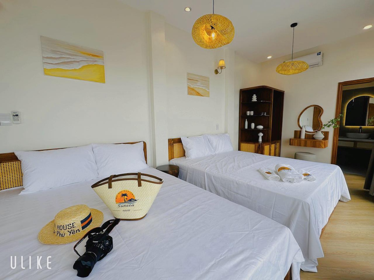 The spacious room features two large beds with fresh linens and minimalist decor. Light fixtures provide soft illumination, while coastal-themed artwork decorates the walls. A wooden shelf holds various decorative items, and a mirror enhances the open feel of the area.
