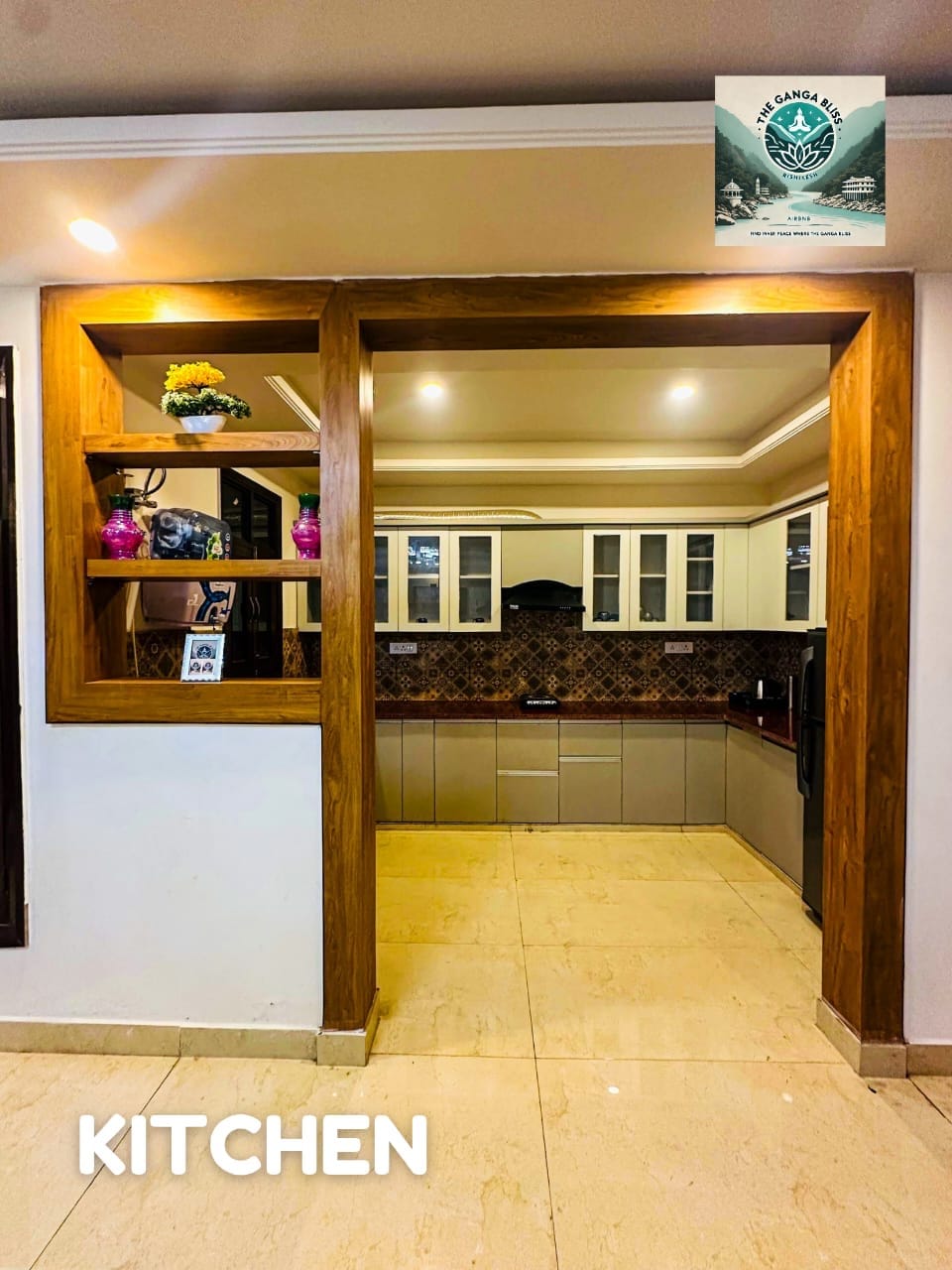 A well-lit kitchen area is visible, featuring modern cabinetry and equipped countertops. Open shelving displays decorative items and plants, adding a homely touch. The space is designed with ample storage, seamlessly connecting to the surrounding living area.
