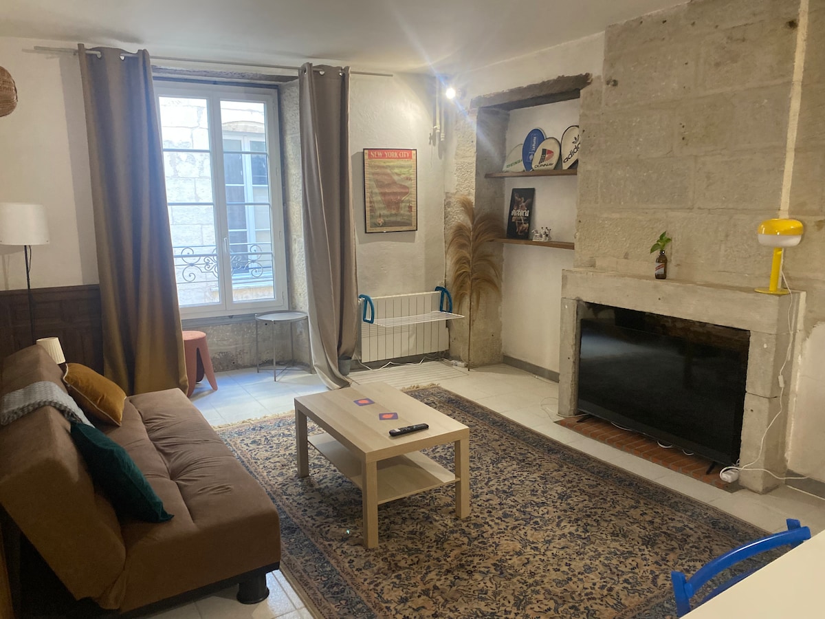 A spacious living area features a comfortable sofa and a low coffee table, positioned on a classic rug. Natural light enters through a window with sheer curtains. A TV is mounted against a stone wall, with decorative shelves showcasing various items.