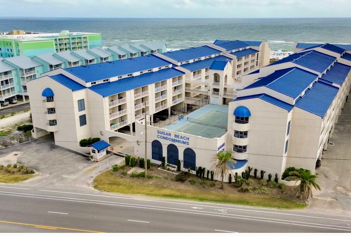 The Sugar Beach Condominiums complex is depicted from an aerial view, showcasing multiple two- and three-story buildings with blue roofs. Lush palm trees and landscaping are visible along the ground level, while the Gulf of Mexico can be seen in the background.