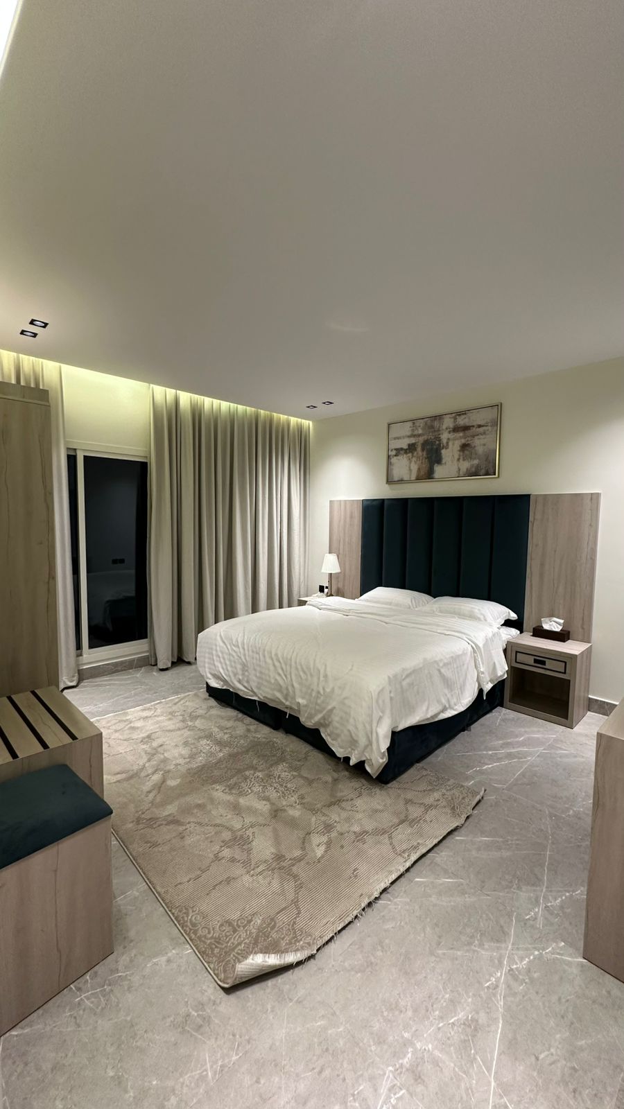 A modern bedroom is featured with a spacious bed dressed in white linens against a dark upholstered headboard. Soft lighting highlights the room's sleek design, while curtains frame the window. Nightstands with lamps are positioned on both sides of the bed, and a rug adds warmth to the floor.