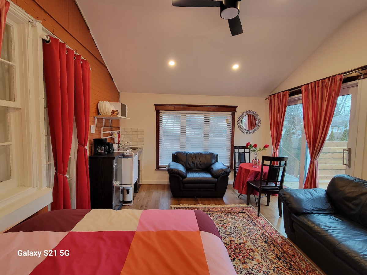 A bright studio features a spacious bed with a contrast of warm colors against the wooden flooring. Cozy seating arrangements include a dark leather couch and a dining table set for two, surrounded by large windows covered with light curtains.