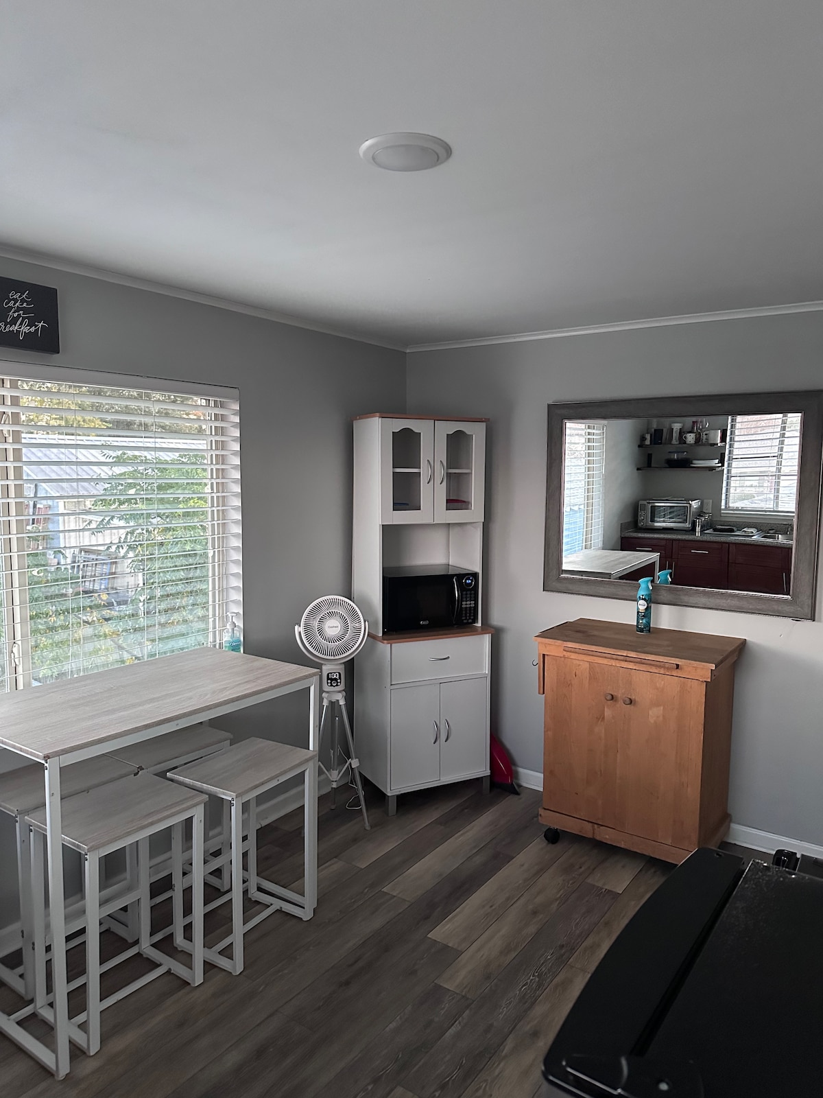 A cozy kitchenette area features a small table with four stools next to a window, allowing natural light to illuminate the space. A microwave and fan are visible on the countertop, with cabinets providing storage. A mirror reflects the inviting ambiance.