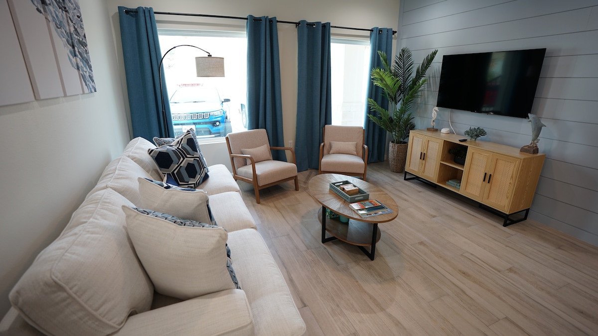 A cozy living room area features a light-colored sectional couch complemented by two armchairs. A medium-sized coffee table is positioned in front of the seating arrangement. Natural light enters through large windows adorned with blue curtains, while a potted plant adds a touch of greenery.