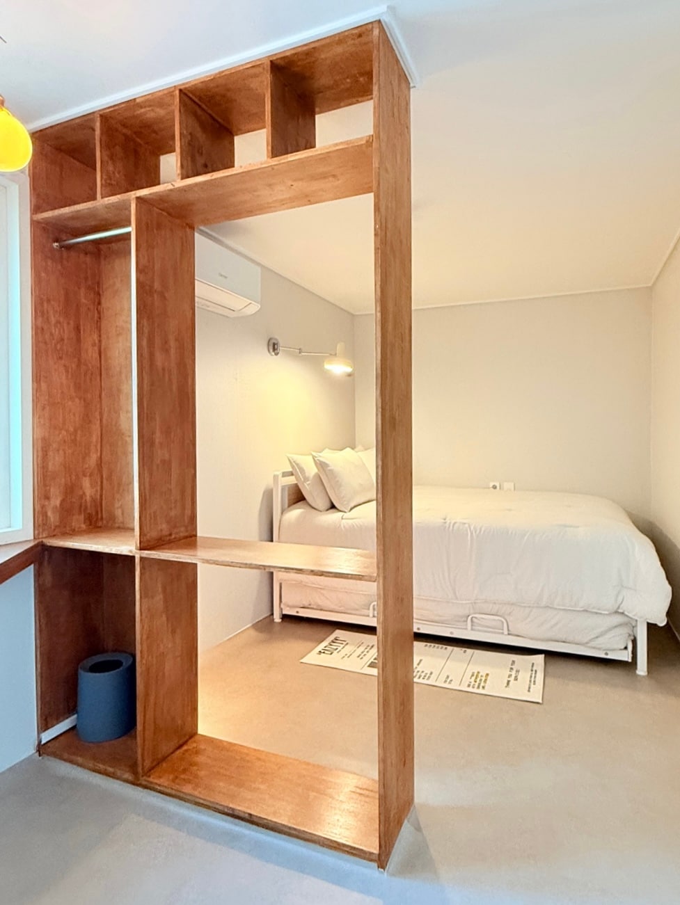 A divided space features an open shelf unit made of warm wood, separating a sleeping area from the rest of the room. The sleeping area includes a spacious bed with neatly arranged white bedding. Soft, natural lighting enhances the calm and inviting atmosphere.