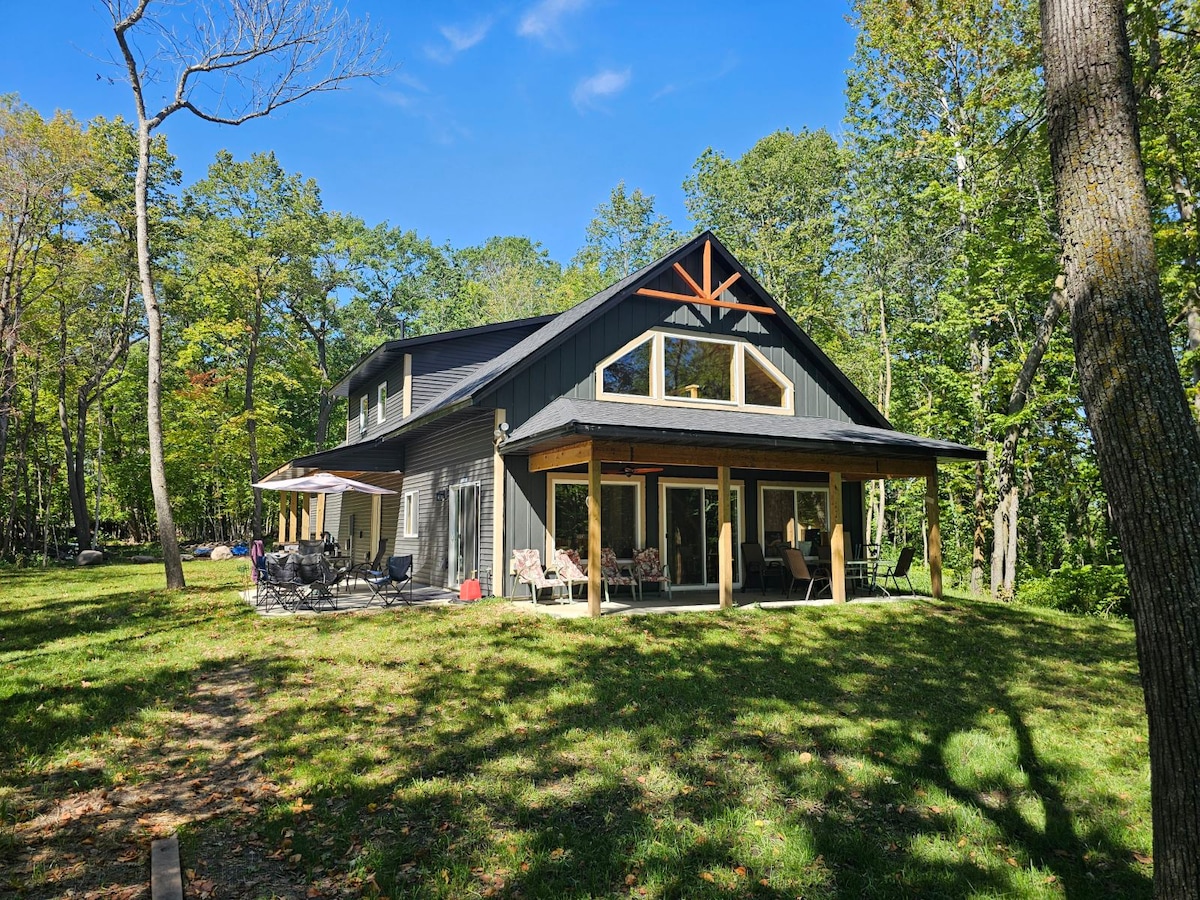 A newly built cabin is depicted, surrounded by lush greenery. Large windows allow natural light to fill the interior. A spacious covered porch features several seating arrangements, providing a relaxing outdoor space. The cabin design incorporates a gable roof with decorative wooden accents.