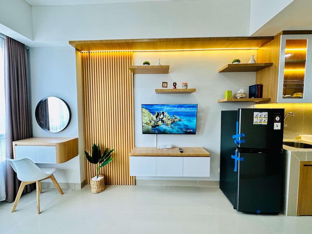 A modern living area is presented with sleek wood accents and a mounted television. A round mirror is positioned above a desk with storage, while a small plant adds a touch of greenery. A black refrigerator is visible nearby, complementing the kitchen design.