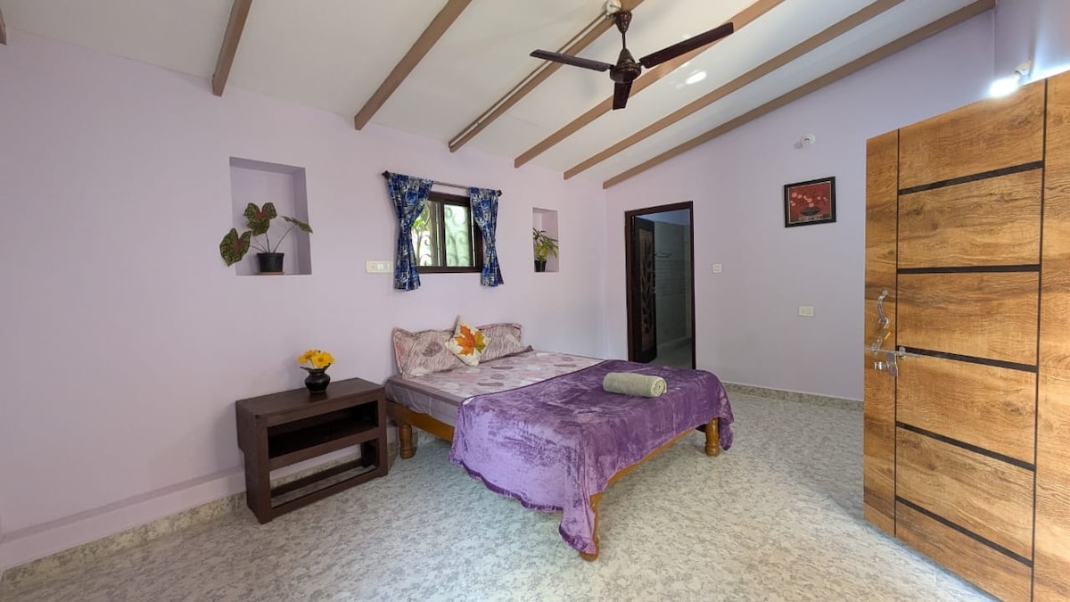 A spacious sleeping area features a double bed covered with a purple bedspread. Natural light enters through a window adorned with blue curtains. The room is complemented by wooden furniture and light purple walls, creating a calming ambiance. A fan is mounted on the ceiling.