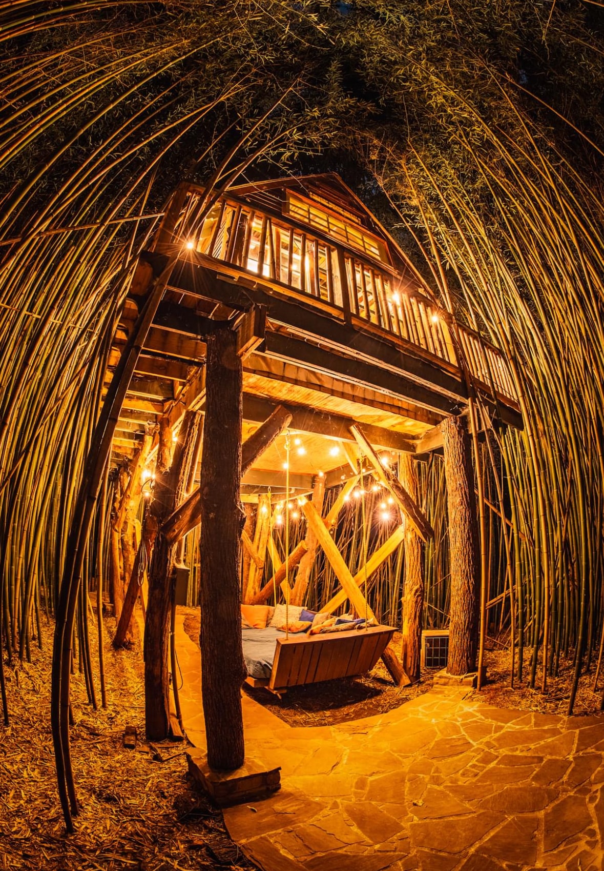 The treehouse is nestled among tall bamboo stalks, illuminated by warm, hanging lights. Its sturdy wooden structure is supported by thick posts, while a swinging rope bed is visible beneath the deck. Natural stone path leads to the inviting entryway.