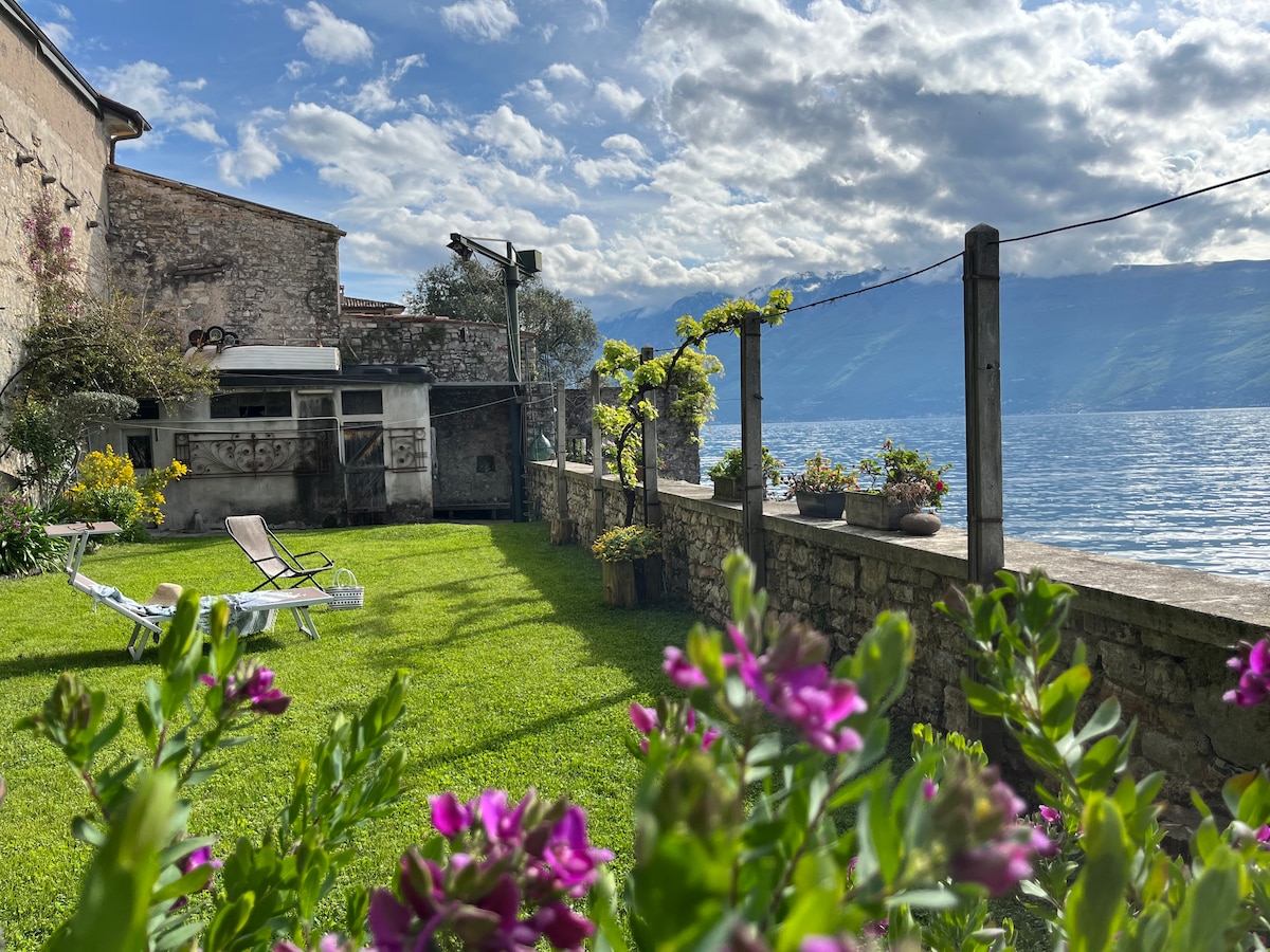 A lush green garden stretches toward the tranquil lake, bordered by a stone wall and adorned with flowering plants. Sun loungers are positioned on the grass, providing a serene spot for relaxation. The expansive lake and mountains are visible in the background, complemented by a vivid sky.