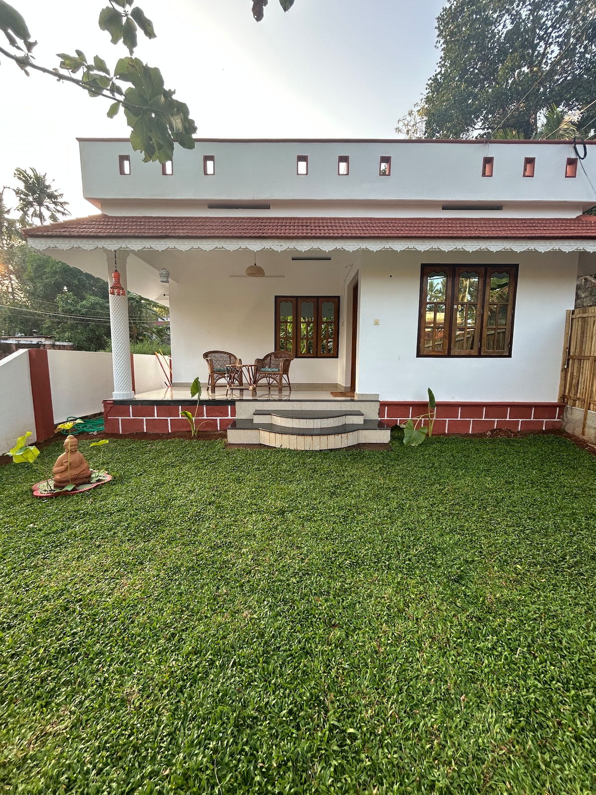 The exterior of the home showcases a charming porch with two wicker chairs, nestled amid a well-kept grassy lawn. The structure features a clean design with large windows, allowing natural light to brighten the space, set against a backdrop of trees for a serene atmosphere.