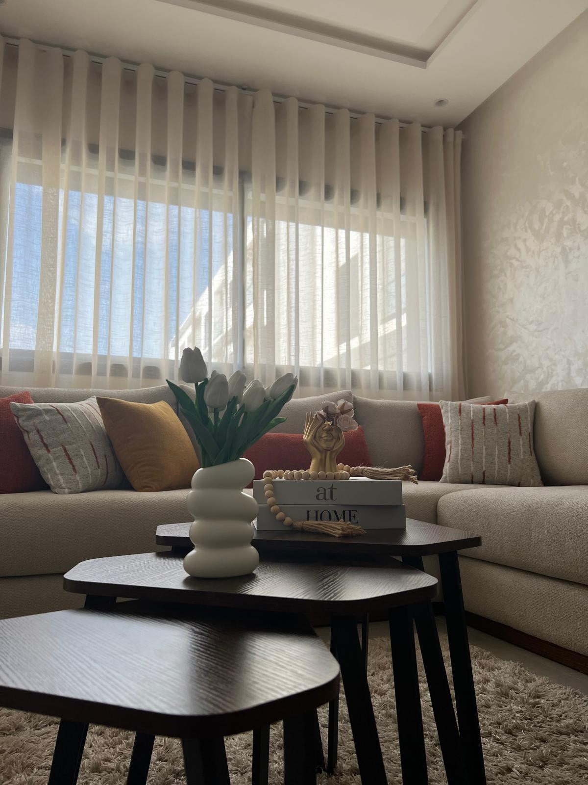 A cozy living area is presented, featuring a light-colored sofa adorned with patterned cushions. Small black tables are arranged at the front, with a decorative vase of white tulips and books stacked on top. Natural light filters through sheer curtains, illuminating the space.