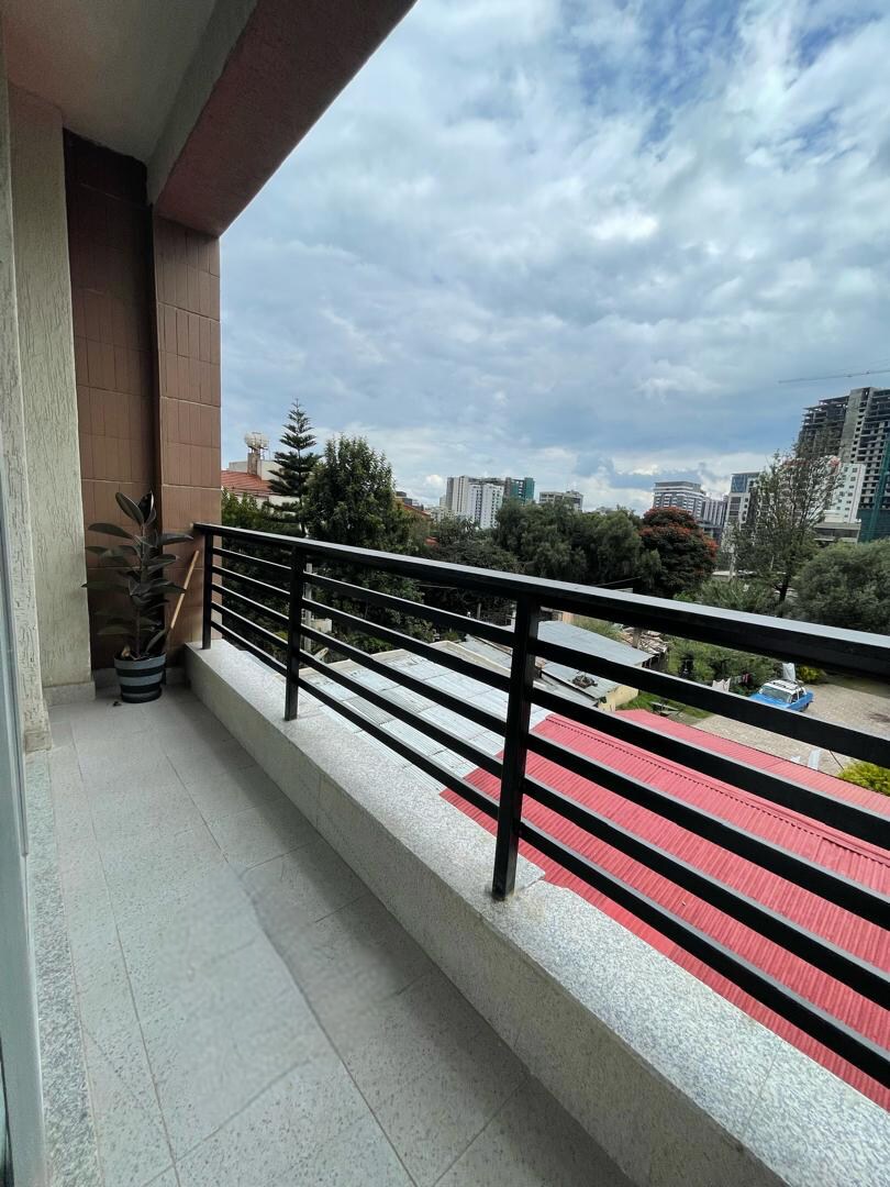 A balcony is shown with a railing overlooking a green landscape and city buildings. A potted plant stands to the side, and the sky features a mix of clouds, adding to the serene atmosphere of the outdoor space.