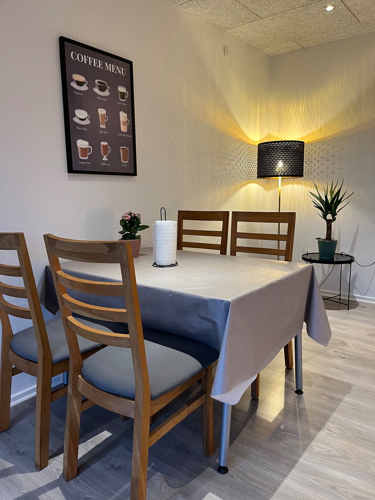 A dining area is presented with a small table covered by a light tablecloth. Four wooden chairs surround the table, and a coffee menu is displayed on the wall. A lamp and a plant provide soft lighting and a touch of greenery.