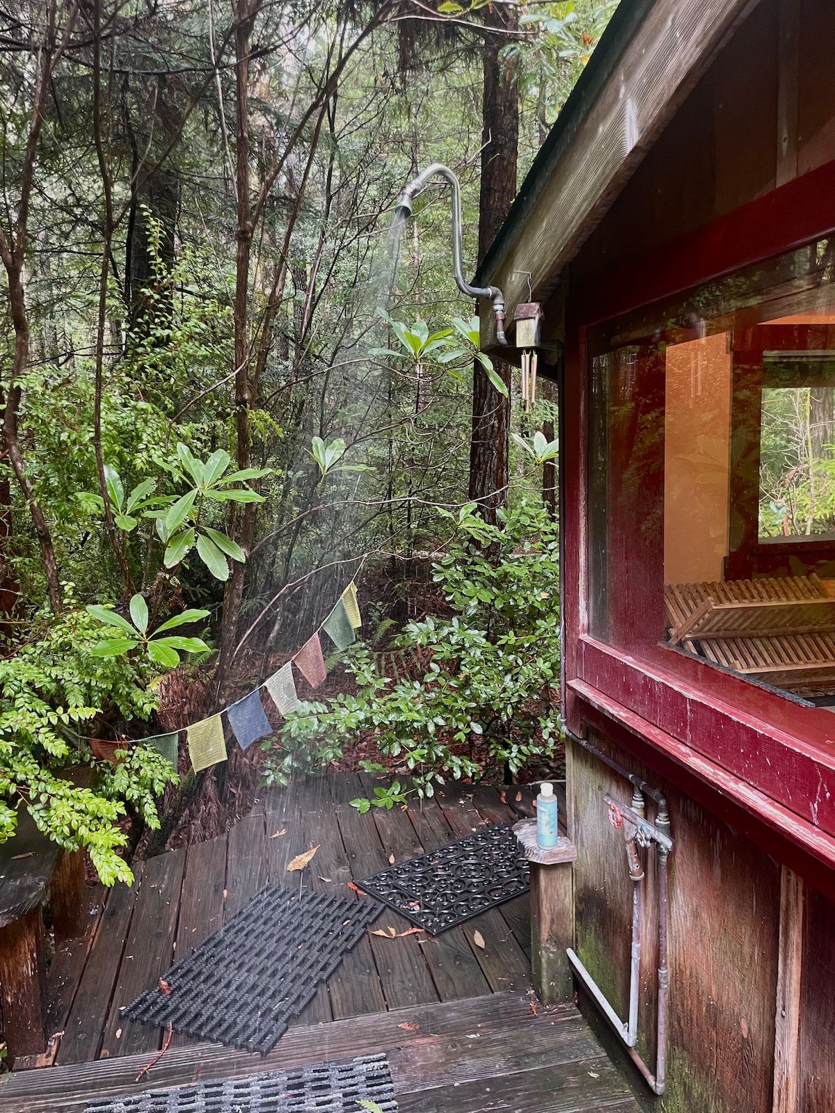 An outdoor shower area is tucked among lush greenery, with wooden decking providing a natural feel. Colorful prayer flags flutter gently in the breeze, adding a touch of color against the backdrop of rich foliage. The red trim of the nearby cottage is visible through the window.